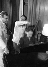 Portada:Plano general de Gregor Piatigorsky observando a Arthur Rubinstein sentado al piano, detrás Jascha Heifetz charlando