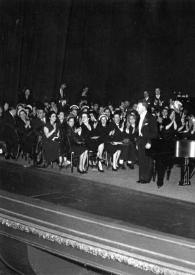Portada:Plano general de Arthur Rubinstein rodeado de espectadores en el escenario saludando, de pie, junto al piano. Fotografía tomada desde el fondo de la sala de conciertos