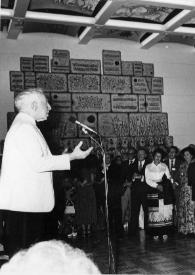 Portada:Plano general de Arthur Rubinstein (perfil derecho) hablando a través de un micrófono hacia un público que se encuentra frente a él