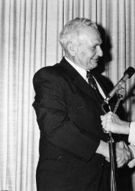 Portada:Plano medio de un hombre y Arthur Rubinstein (perfil izquierdo) hablando por un micrófono y saludándose