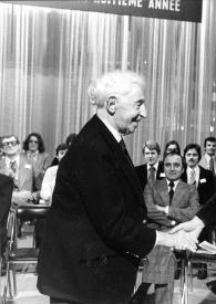 Portada:Plano medio de Arthur Rubinstein (perfil derecho) estrechando la mano a un hombre (perfil izquierdo) con un diploma en la mano, detrás los demás participantes les observan.