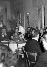 Portada:Plano medio de Arthur Rubinstein de pie entre las mesas con la copa alzada, hablando a los asistentes, entre ellos sentados en diferentes mesas: Sol Hurok