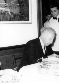 Portada:Plano medio de Arthur Rubinstein firmando un autógrafo a un hombre sentado en una mesa, detrás un camarero del restaurante les observa